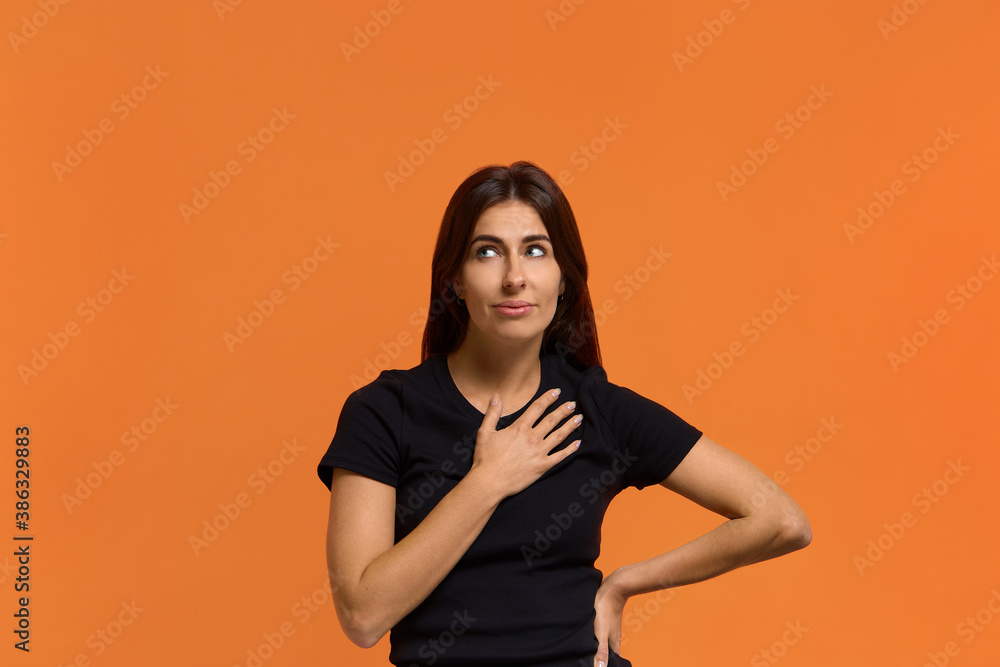 Obraz premium Very unexpected. Portrait of happy caucasian woman in black t-shirt folds arm on chest, expresses gratitude, promises something, has tender smile and long hair. Isolated over an orange wall