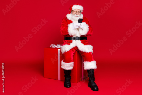 Full length body size view of his he handsome cheerful bearded Santa father wearing warm coat sitting on box use device app delivery celebrate isolated bright vivid shine vibrant red color background