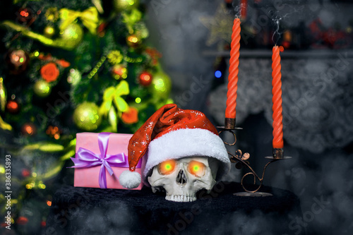 Fotografie Human skull with evil glowing eyes in a Christmas hat lies in fog on a table next to smoking candles and a gift against the background of a decorated Christmas tree