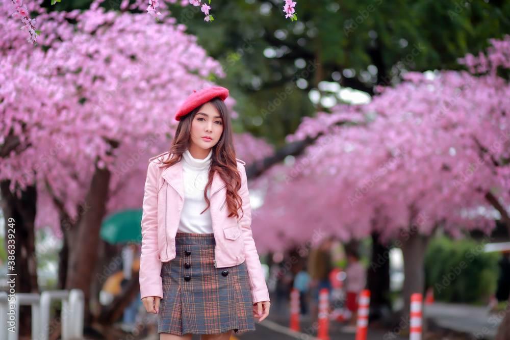 A traveler hipster woman sightseeing wear a red hat and a smooth leather dress with beautiful sakura cherry blossoms tree full blooming in pink color in the park on a spring day.