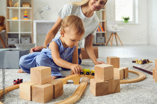 Young nanny and toddler boy playing with wooden blocks in modern cozy nursery room