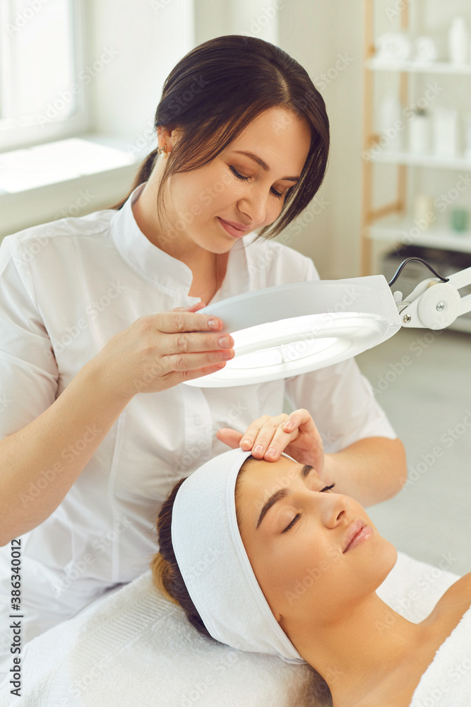 Woman dermatologist examining womans facial skin elasticity and condition under lamp Woman dermatologist examining womans facial skin elasticity and condition under lamp