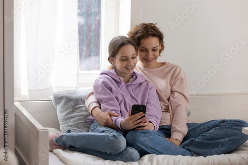 Smiling Caucasian loving mother and teen daughter sit relax on bed at home use smartphone together. Happy mom and teenage girl child have fun rest watch video on cellphone gadget. Technology concept.