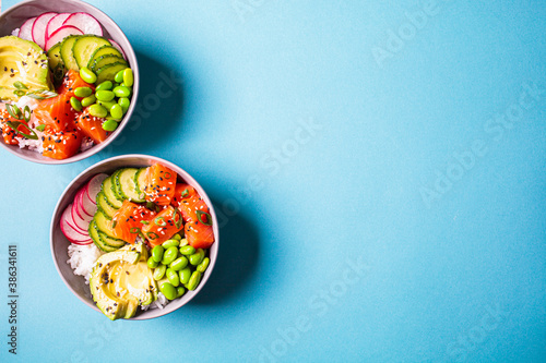 Poke bowl with salmon, rice, avocado, edamame beans, cucumber and radish in gray bowls, top view. Hawaiian ahi poke bowl, blue background.