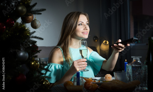 Portrait of smiling young woman drinking champagne and watching TV at Christmas night