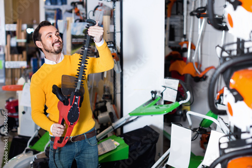 Young happy cheerful positive man choosing hedge cutter in garden equipment shop