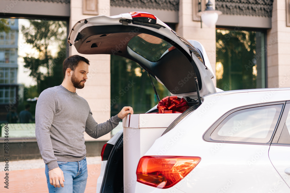Naklejka premium Bearded young delivery man standing near car, large festive white box with beautiful red bow at truck. Courier preparing to take parcel out of trunk of car and deliver it to customer.