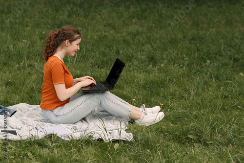 Young smiling girl working with laptop and sitting on the green grass in sunny weather city park or yard. Happy woman in the city park with copy space.