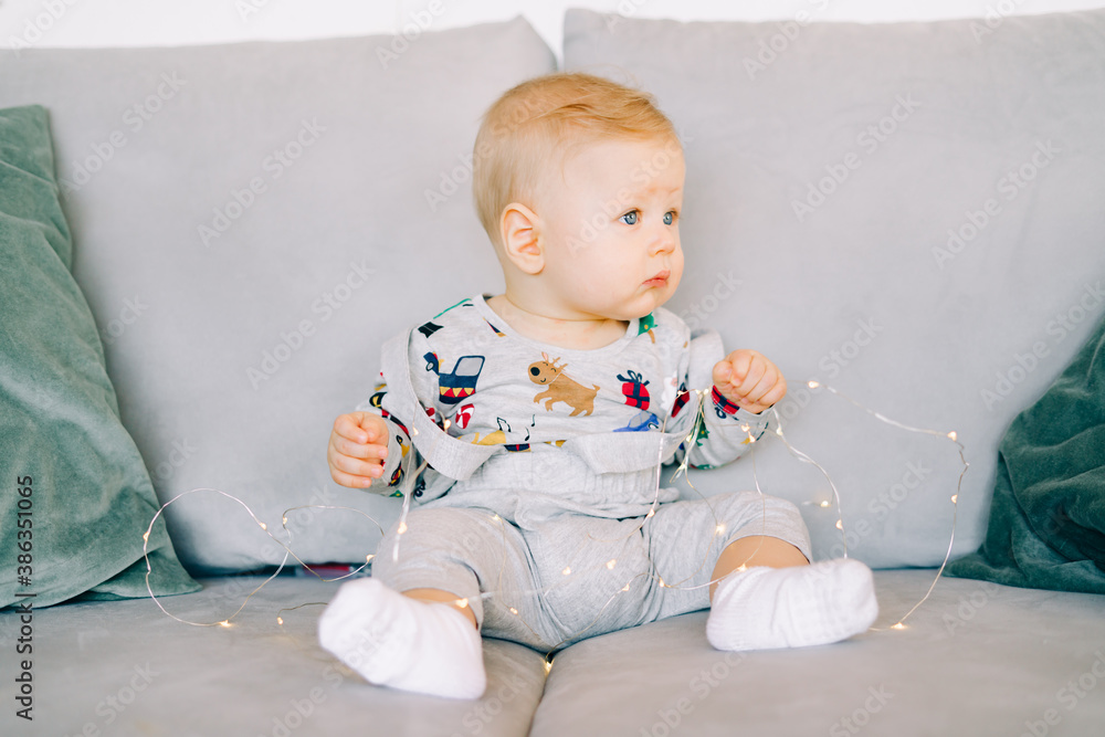 a cute little boy who is teething is sitting in a festive Christmas costume on a gray sofa and playing with a bright light garland.Christmas concept a cute little boy who is teething is sitting in a festive Christmas costume on a gray sofa and playing with a bright light garland.Christmas concept