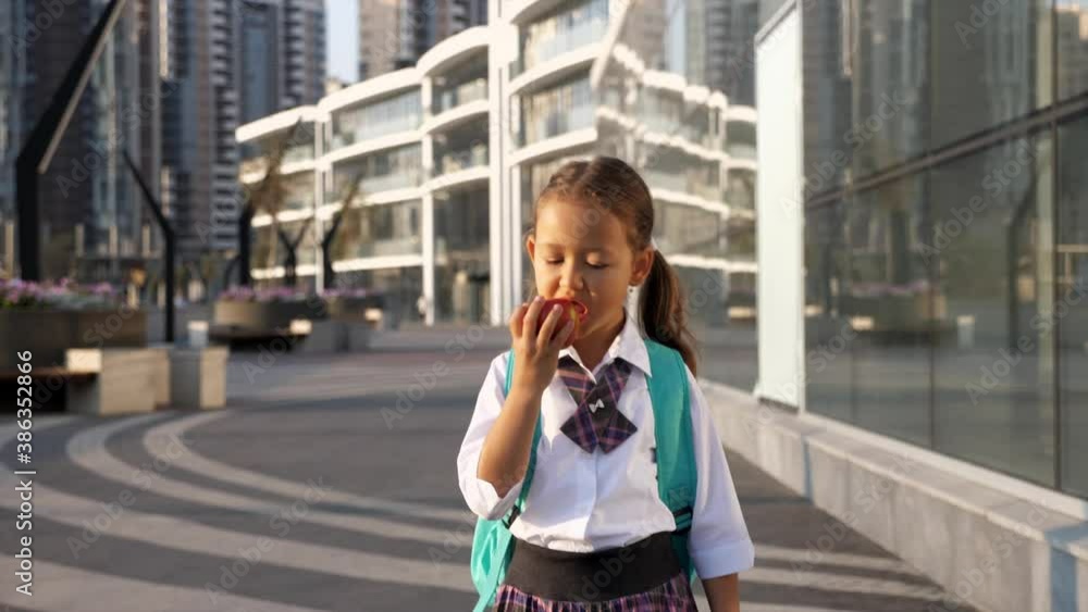Little child girl with a backpack in school uniform is going to school in city landscape and eating the apple, front view. Primary school pupil is going to study, steadicam shot. Little child girl with a backpack in school uniform is going to school in city landscape and eating the apple, front view. Primary school pupil is going to study, steadicam shot.