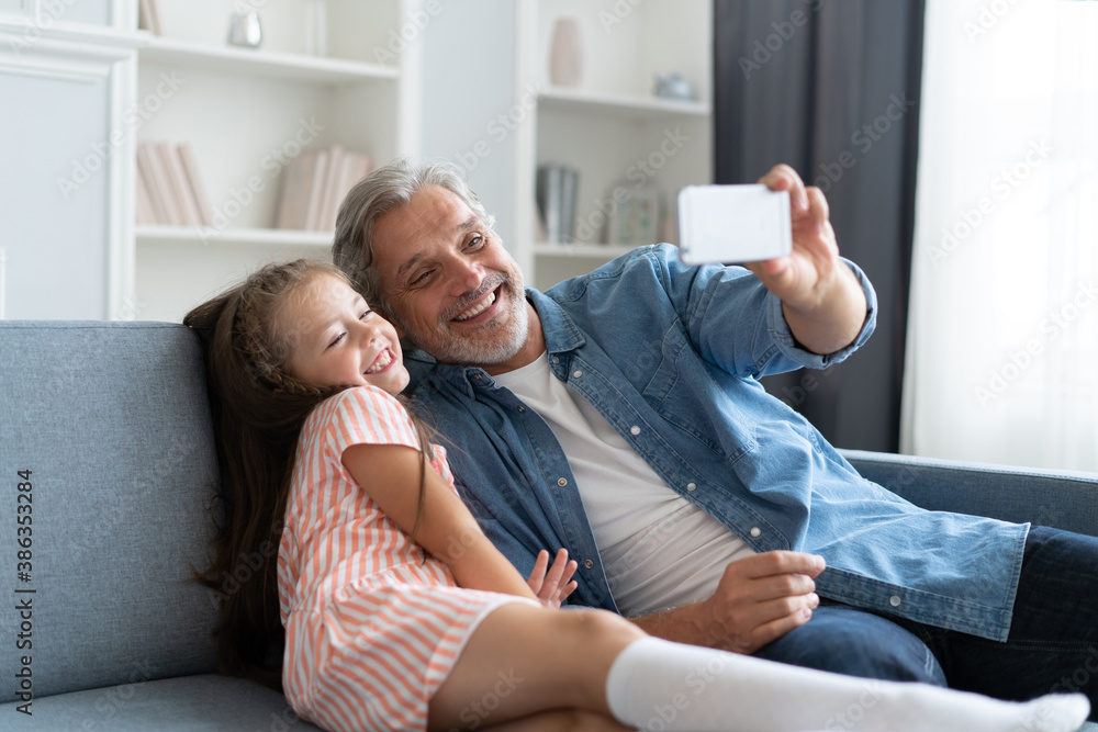Fototapeta premium Funny selfie with dad. Self portrait of father and his little daughter taking selfie while sitting on the sofa