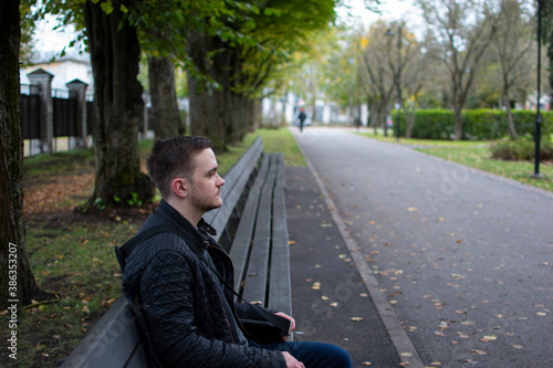 young adult guy sits on a park bench in dark clothes, green park autumn