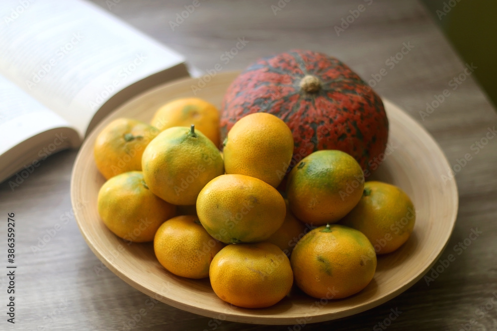 Wooden bowl with tangerines and a pumpkin and open book on a table. Selective focus. Wooden bowl with tangerines and a pumpkin and open book on a table. Selective focus.