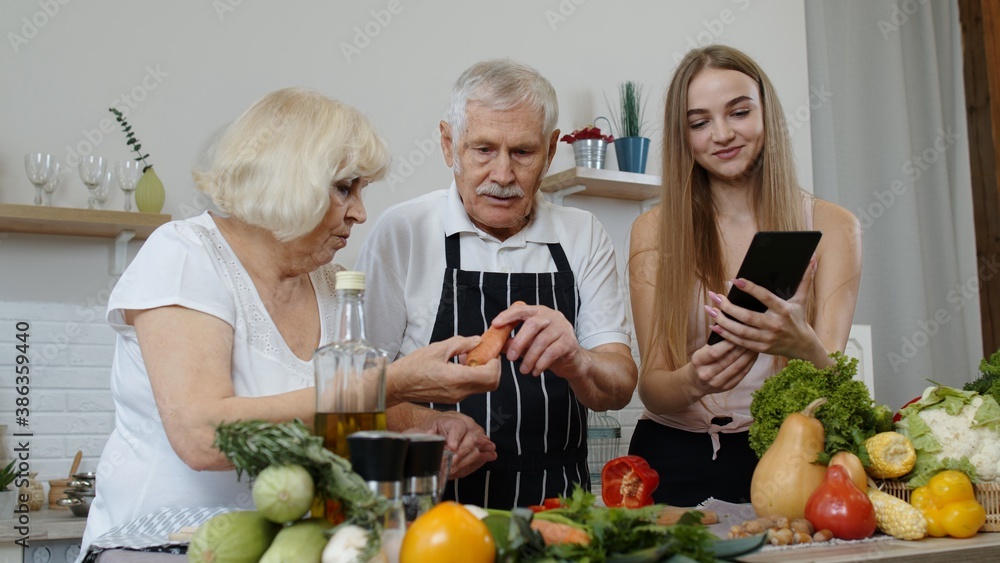 Mature grandparents couple cutting vegetables for salad, listening recipe from girl with tablet