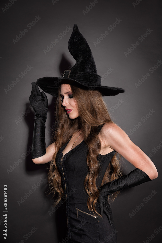 Witch woman with long curly hair and perfect makeup and red lips in witches black hat. Green background, copy space. Halloween costume