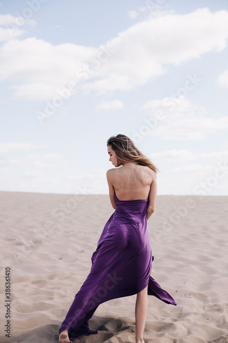 Fototapeta Naklejka Na Ścianę i Meble -  A young, slender girl in a beige dress with purple cloth in her hands posing in the desert in the wind