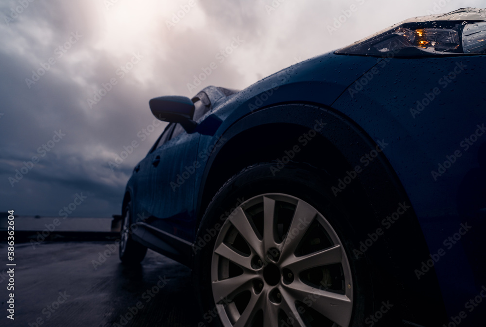New luxury blue SUV car parked on concrete road beside the beach on rainy day with stormy sky. Front view of blue SUV car with sport design. Raindrops on car. Road trip travel. Driving in bad weather.