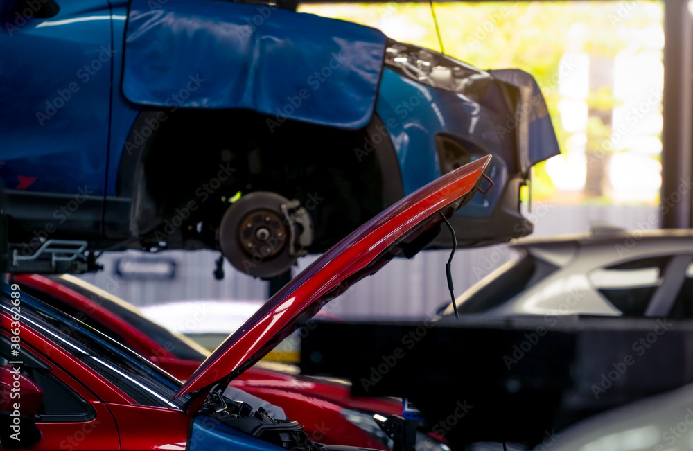 Auto repair shop. Red car opened hood on blurred background of blue SUV car lifted in the garage for repair and maintenance service. Car check-up at service station. Car body lifted in workshop. Auto repair shop. Red car opened hood on blurred background of blue SUV car lifted in the garage for repair and maintenance service. Car check-up at service station. Car body lifted in workshop.