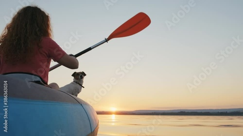 Happy young woman paddling on an inflatable kayak with her dog Jack Russell Terrier on water of sea against beautiful orange sunset slow motion. Family Sports. Summer. Travel. Tourism. Relax. Freedom