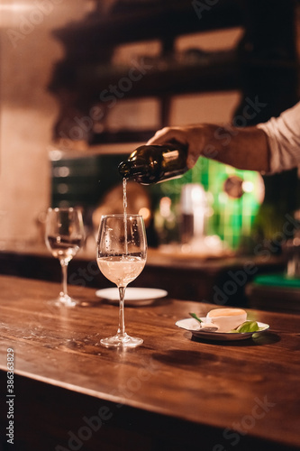 A barman pouring white wine into a glass. Wooden table in a dark bar © Надежда Филатова