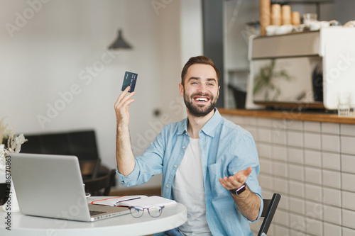 Smiling young man sitting alone at table in coffee shop cafe restaurant indoors working studying on laptop computer rising hand with credit bank card for pay. Freelance mobile office business concept.