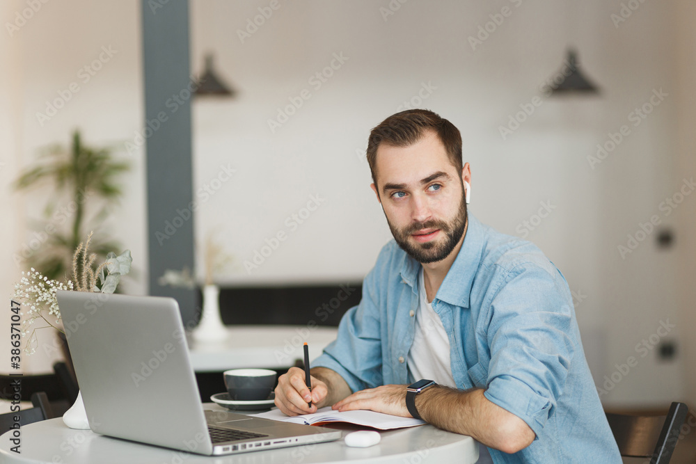 Serious young man in air pods sitting alone at table in coffee shop cafe restaurant indoors working or studying on laptop pc computer writing in notebook. Freelance mobile office business concept. Serious young man in air pods sitting alone at table in coffee shop cafe restaurant indoors working or studying on laptop pc computer writing in notebook. Freelance mobile office business concept.