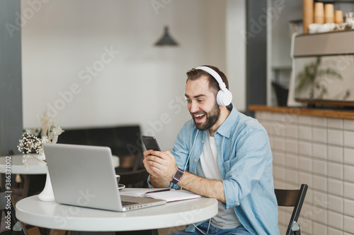 Excited young man sitting at table in coffee shop cafe restaurant indoors working studying on laptop pc computer listening music with headphones cell phone. Freelance mobile office business concept.