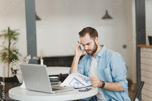 Preoccupied puzzled young man sitting alone at table in coffee shop cafe restaurant indoors working or studying on laptop pc computer with papers document. Freelance mobile office business concept.