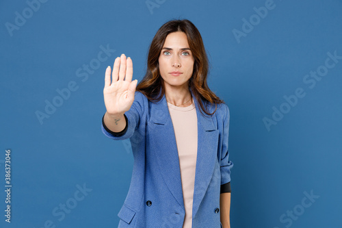 Serious confident attractive young brunette woman 20s wearing basic jacket standing showing stop gesture with palm looking camera isolated on bright blue colour wall background, studio portrait.