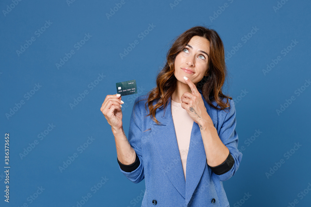 Pensive pretty young brunette woman 20s wearing basic jacket standing holding in hand gift certificate put hand prop up on chin looking up isolated on bright blue colour background studio portrait. Pensive pretty young brunette woman 20s wearing basic jacket standing holding in hand gift certificate put hand prop up on chin looking up isolated on bright blue colour background studio portrait.