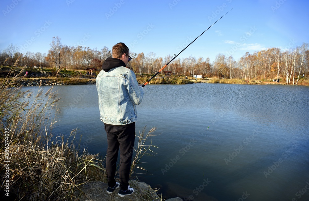 Young fisherman in denim jacket standing on stone, fishing in the lake. he holding the rod and looking at float against autumn forest in park. Young fisherman in denim jacket standing on stone, fishing in the lake. he holding the rod and looking at float against autumn forest in park.