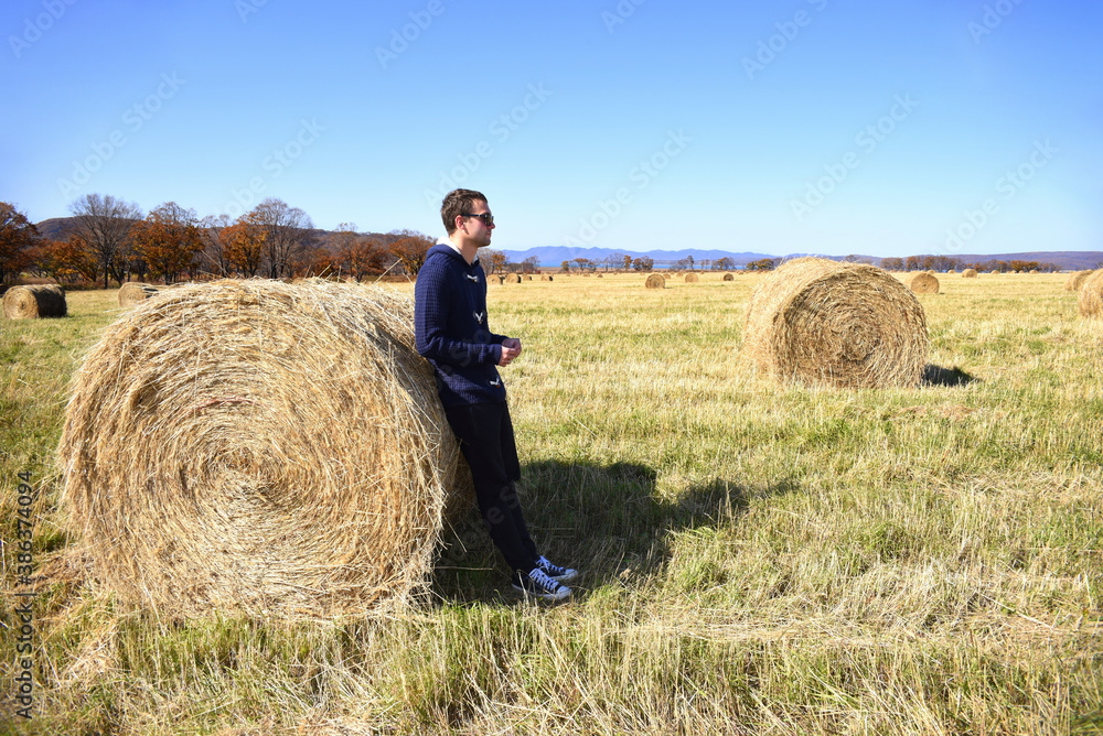 Young brunette man in warm jacket leaning against haystack in field with bales of hay. he looking to the right. Primorsky kray, Russia. Beauty in nature. Copy space Young brunette man in warm jacket leaning against haystack in field with bales of hay. he looking to the right. Primorsky kray, Russia. Beauty in nature. Copy space