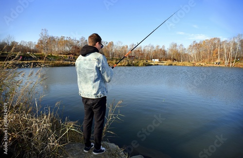 Young fisherman in denim jacket standing on stone, fishing in the lake. he holding the rod and looking at float against autumn forest in park. 
