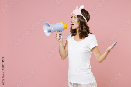 Funny young woman in pajamas home wear sleep mask screaming in megaphone spreading hands looking aside resting at home isolated on pink background studio portrait. Relax good mood lifestyle concept.