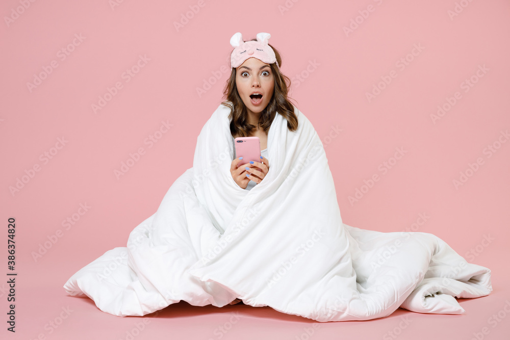 Amazed young woman in pajamas home wear sleep mask sit using mobile cell phone keeping mouth open resting at home isolated on pastel pink background studio portrait. Relax good mood lifestyle concept. Amazed young woman in pajamas home wear sleep mask sit using mobile cell phone keeping mouth open resting at home isolated on pastel pink background studio portrait. Relax good mood lifestyle concept.