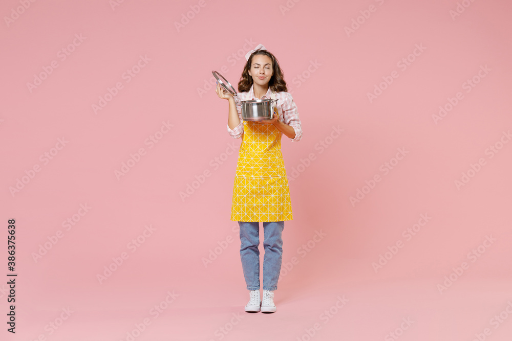 Full length portrait of smiling young woman housewife in apron hold saucepan with lid feeling food smell while doing housework isolated on pastel pink colour background studio. Housekeeping concept. - obrazy, fototapety, plakaty 