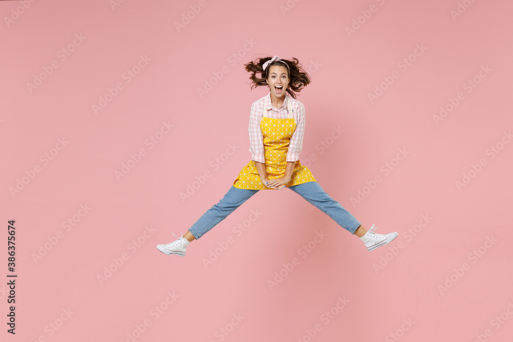 Full length portrait of excited young brunette woman housewife 20s in yellow apron jumping spreading legs while doing housework isolated on pastel pink colour background studio. Housekeeping concept. Full length portrait of excited young brunette woman housewife 20s in yellow apron jumping spreading legs while doing housework isolated on pastel pink colour background studio. Housekeeping concept.