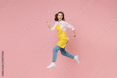 Full length side view portrait of funny young woman housewife 20s in yellow apron jumping like running while doing housework isolated on pastel pink colour background studio. Housekeeping concept.