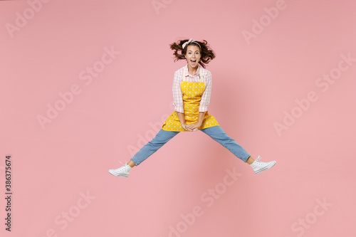 Full length portrait of excited young brunette woman housewife 20s in yellow apron jumping spreading legs while doing housework isolated on pastel pink colour background studio. Housekeeping concept.