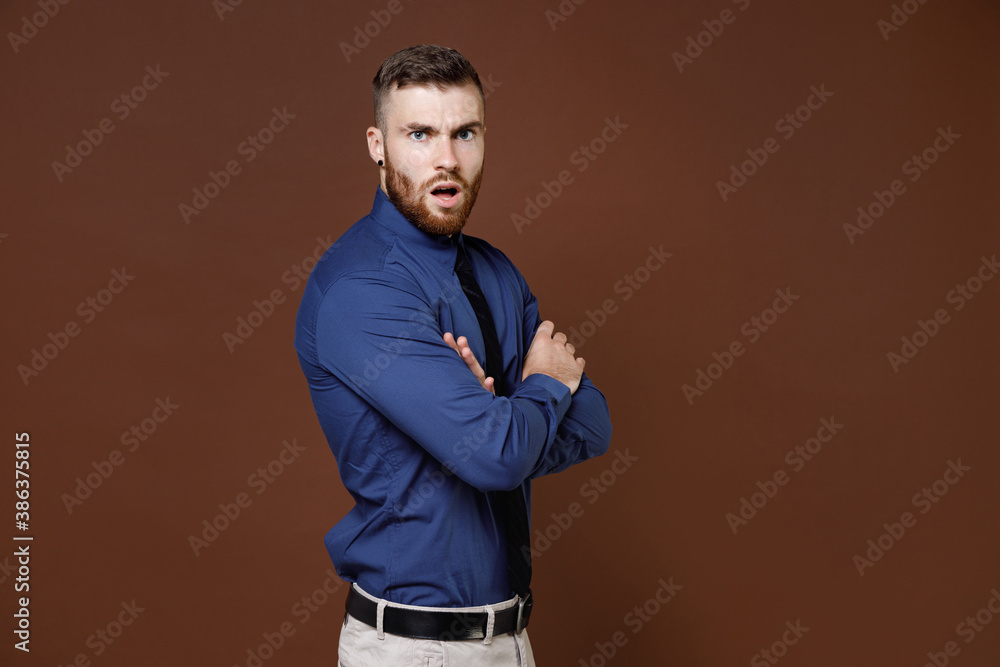 Side view of displeased shocked young business man wearing blue shirt tie holding hands crossed looking camera isolated on brown background studio portrait. Achievement career wealth business concept. Side view of displeased shocked young business man wearing blue shirt tie holding hands crossed looking camera isolated on brown background studio portrait. Achievement career wealth business concept.