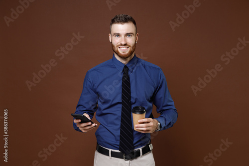Smiling bearded young business man in blue shirt tie using mobile cell phone hold paper cup of coffee or tea isolated on brown background studio portrait. Achievement career wealth business concept.