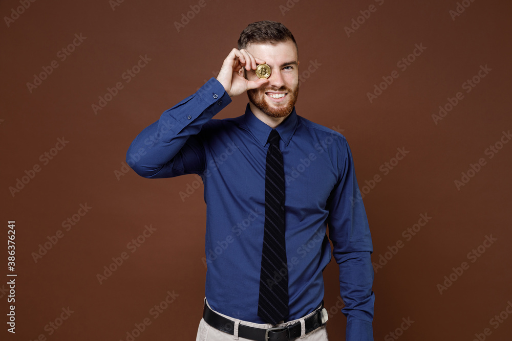 Cheerful bearded young business man wearing blue shirt tie covering eye with bitcoin coin currency isolated on brown colour background studio portrait. Achievement career wealth business concept. Cheerful bearded young business man wearing blue shirt tie covering eye with bitcoin coin currency isolated on brown colour background studio portrait. Achievement career wealth business concept.
