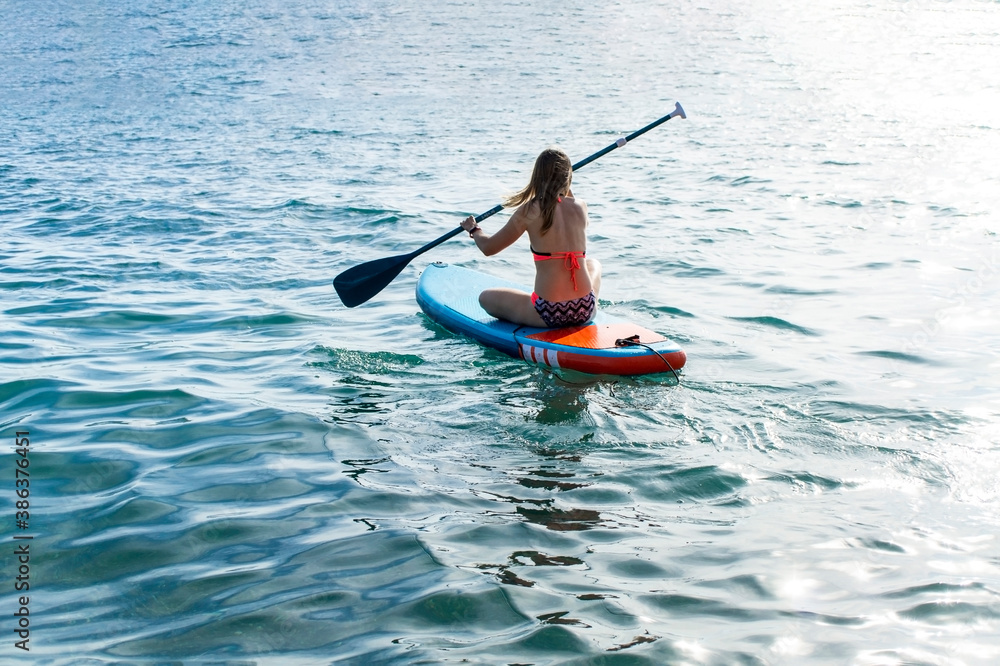 Young blond caucasian woman from the back sitting and swimming on a blue paddle board on the sea. Paddleboarding, activity, sup, pad, water sport Young blond caucasian woman from the back sitting and swimming on a blue paddle board on the sea. Paddleboarding, activity, sup, pad, water sport