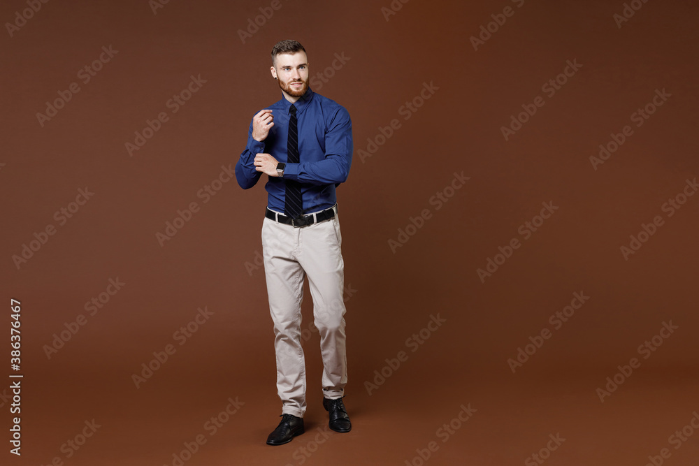 Full length of successful bearded young business man in blue shirt tie straightening sleeves looking aside isolated on brown background studio portrait. Achievement career wealth business concept. Full length of successful bearded young business man in blue shirt tie straightening sleeves looking aside isolated on brown background studio portrait. Achievement career wealth business concept.