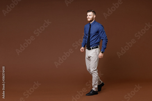 Full length side view of smiling successful young business man wearing blue shirt tie looking aside isolated on brown colour background studio portrait. Achievement career wealth business concept.