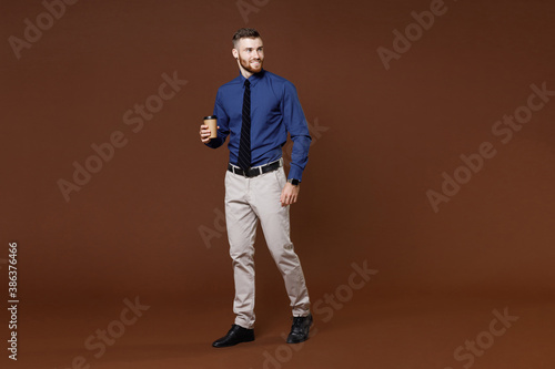 Full length of smiling bearded young business man in blue shirt tie hold paper cup of coffee tea looking aside isolated on brown background studio portrait. Achievement career wealth business concept.