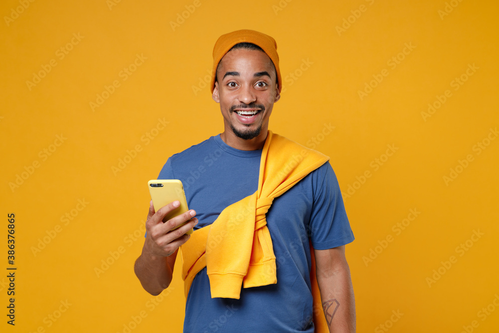 Excited cheerful young african american man 20s wearing basic casual blue t-shirt hat standing using mobile cell phone typing sms message isolated on bright yellow colour background studio portrait.