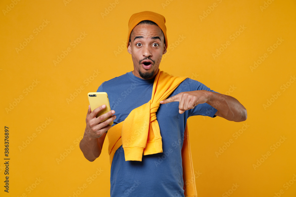 Shocked young african american man 20s in basic casual blue t-shirt hat standing pointing index finger on mobile cell phone typing sms message isolated on bright yellow background studio portrait.