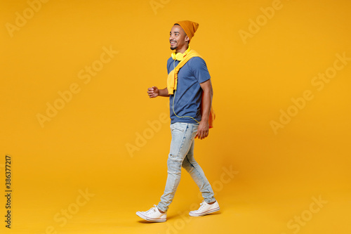 Full length side view of smiling cheerful handsome funny young african american man 20s in blue t-shirt hat walking going looking aside isolated on bright yellow colour background, studio portrait.