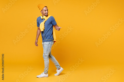 Full length side view of smiling handsome young african american man 20s wearing blue t-shirt hat standing pointing index finger aside isolated on bright yellow colour background, studio portrait.